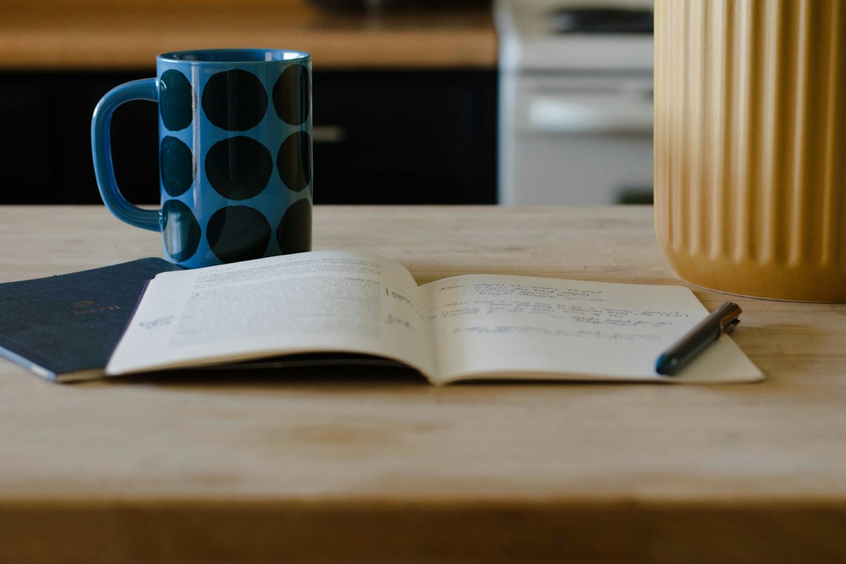 An open journal and pen on a wooden table beside a mug